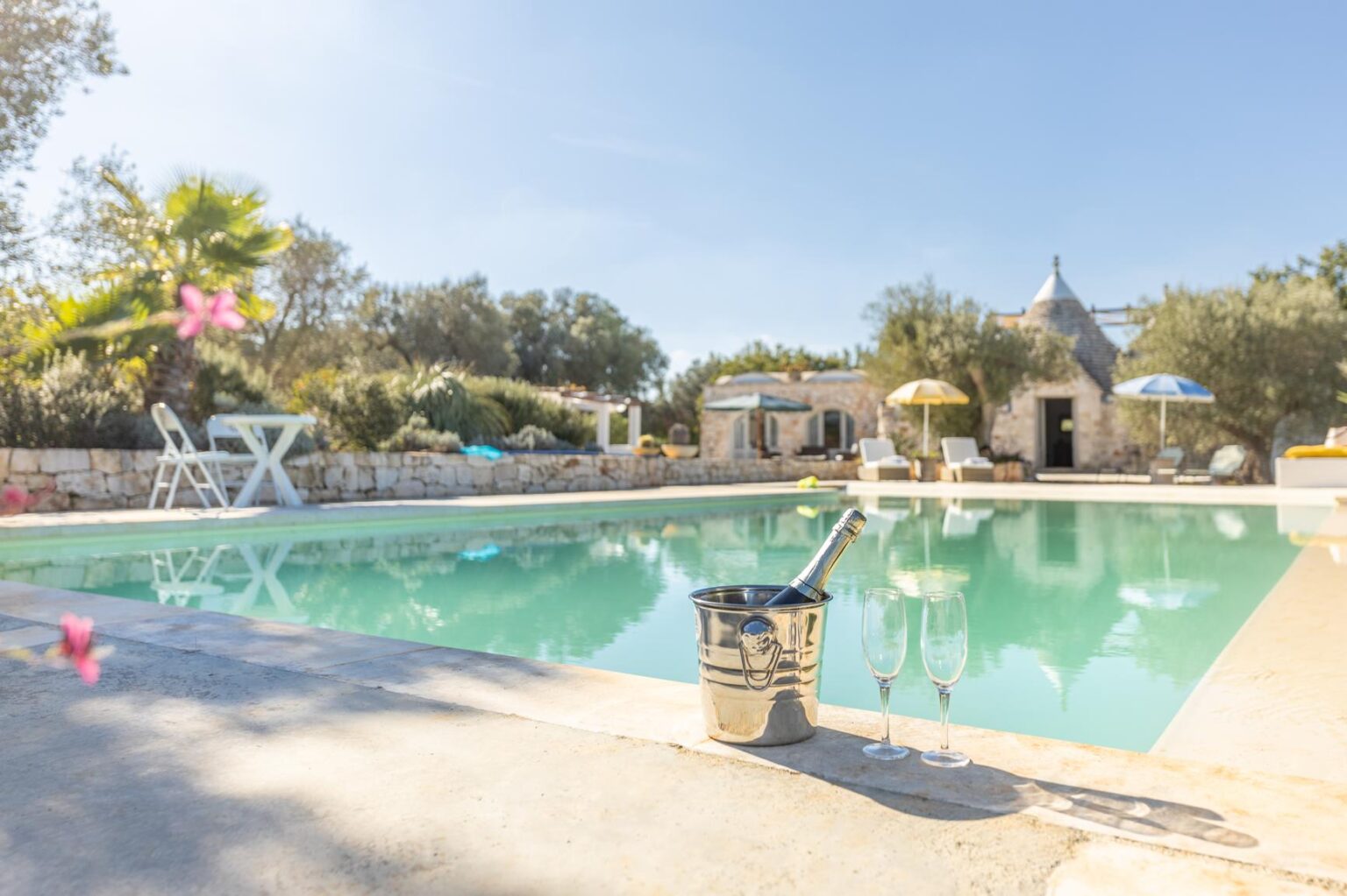 Scenic outdoor pool surrounded by lush greenery and a charming stone structure, featuring a relaxing atmosphere with lounge chairs and colorful umbrellas under a clear blue sky. | Only on SopranoVillas