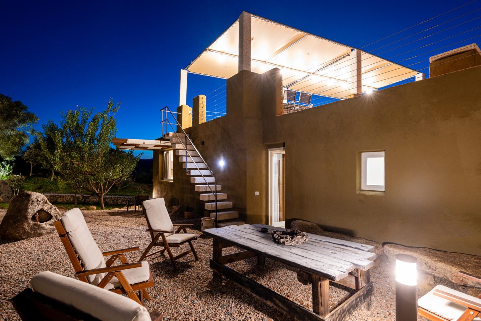 Outdoor seating area with wooden chairs and a table, illuminated at night, featuring a staircase leading to a rooftop deck, surrounded by landscaping and gravel. | Only on SopranoVillas