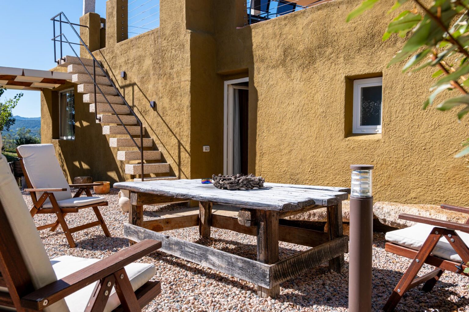 Outdoor patio area featuring a rustic wooden table and chairs, with a staircase leading to an upper level, surrounded by natural landscaping. Ideal for relaxation and entertaining in a serene environment. | Only on SopranoVillas
