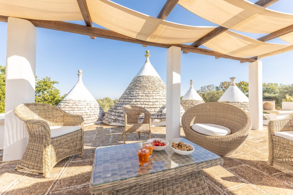 Cozy outdoor seating area with woven furniture overlooking traditional trulli houses, featuring a table with fruit and drinks, under a sunshade in a sunny landscape. | Only on SopranoVillas