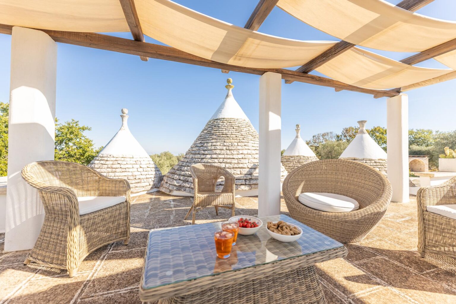 Cozy outdoor seating area with woven furniture overlooking traditional trulli houses, featuring a table with fruit and drinks, under a sunshade in a sunny landscape. | Only on SopranoVillas