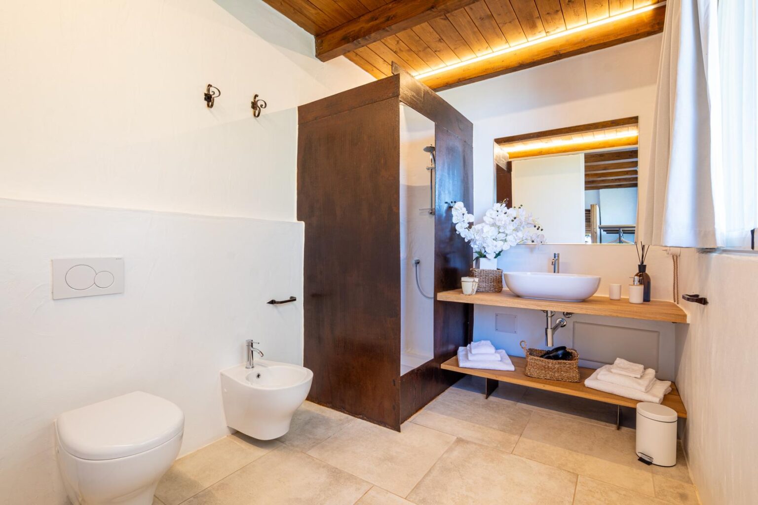 Modern bathroom featuring a walk-in shower, a stylish sink on a wooden countertop, and elegant decor with flowers. The space includes a toilet and bidet, complemented by neutral tile flooring and wooden ceiling beams, creating a serene and inviting atmosphere. | Only on SopranoVillas