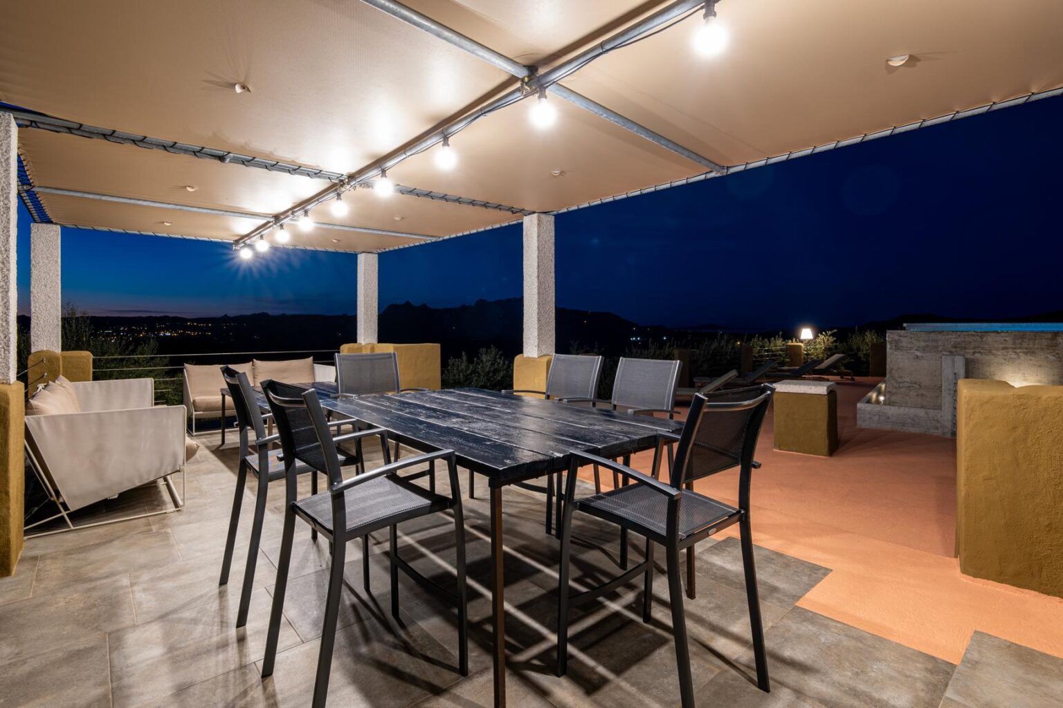 Modern outdoor dining area at night featuring a large black table surrounded by sleek chairs, illuminated by overhead lights, with a scenic view of mountains and city lights in the background. Perfect for evening gatherings and entertaining. | Only on SopranoVillas
