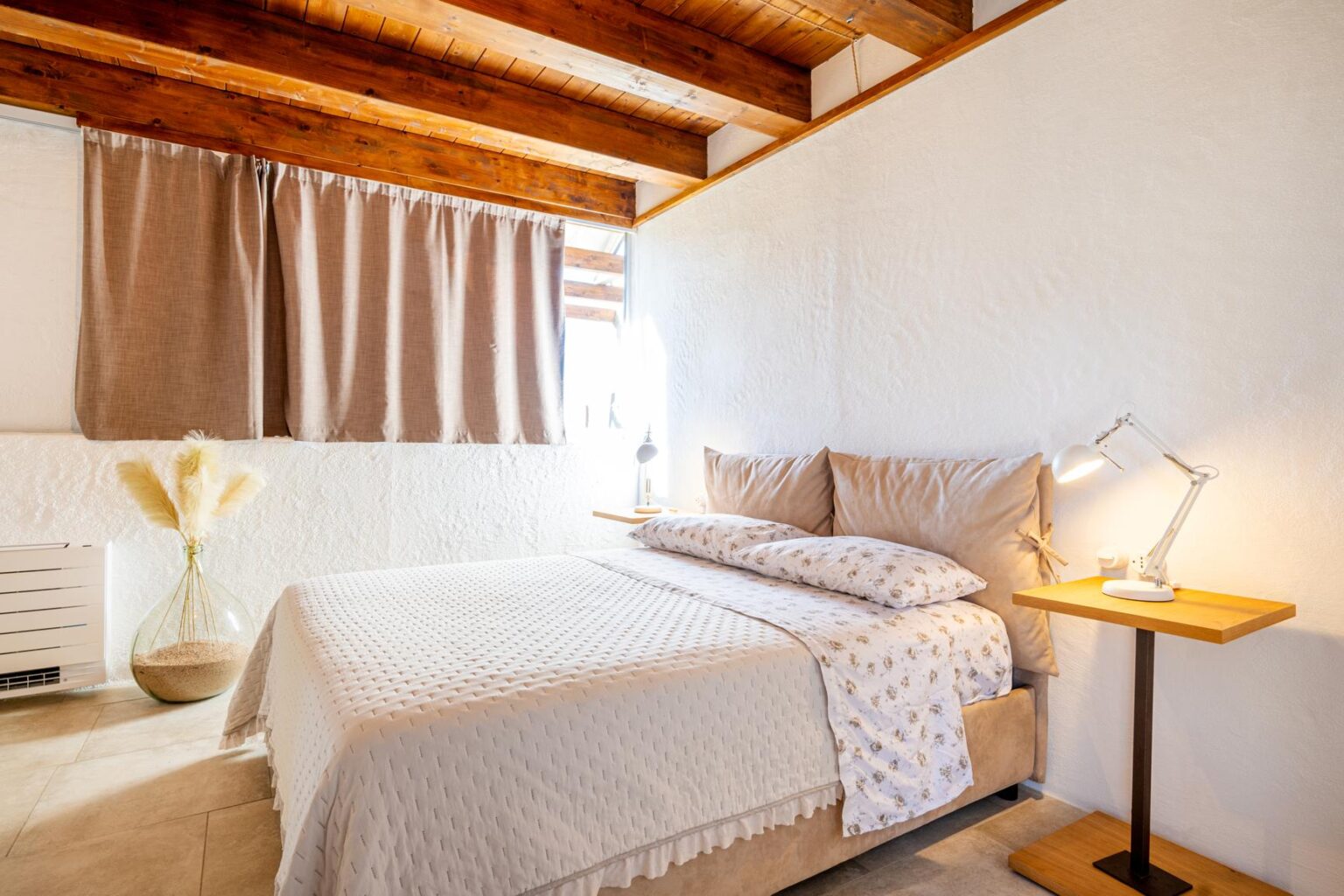 Cozy bedroom featuring a queen-sized bed with soft beige bedding, complemented by a wooden bedside table and a modern desk lamp. Natural light streams through beige curtains, enhancing the warm atmosphere of the room, which includes decorative elements like a glass vase with pampas grass and rustic wooden beams on the ceiling. | Only on SopranoVillas