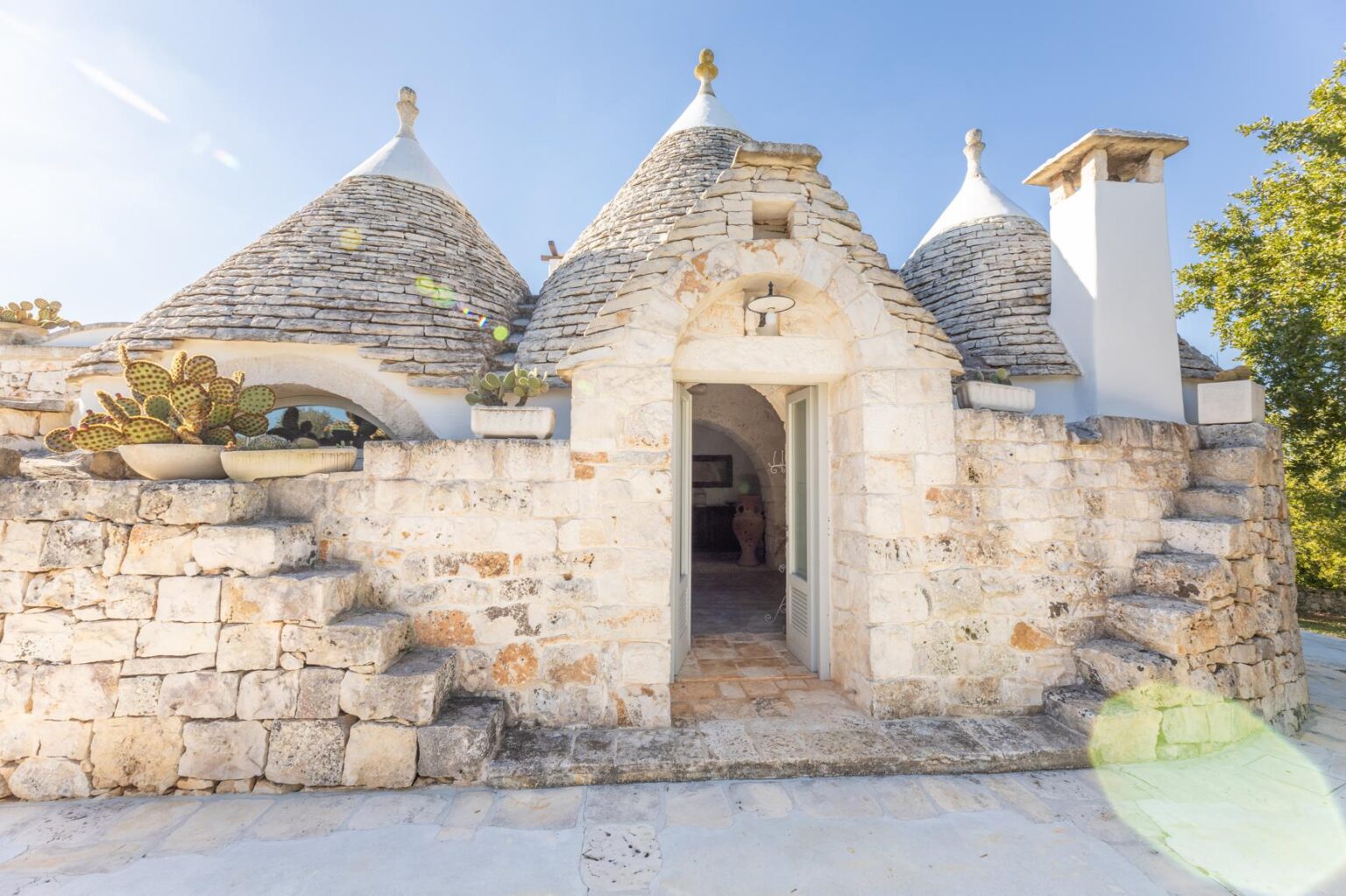 Trullo house in Puglia, Italy, featuring traditional conical roofs and stone walls, surrounded by greenery and a clear blue sky. | Only on SopranoVillas