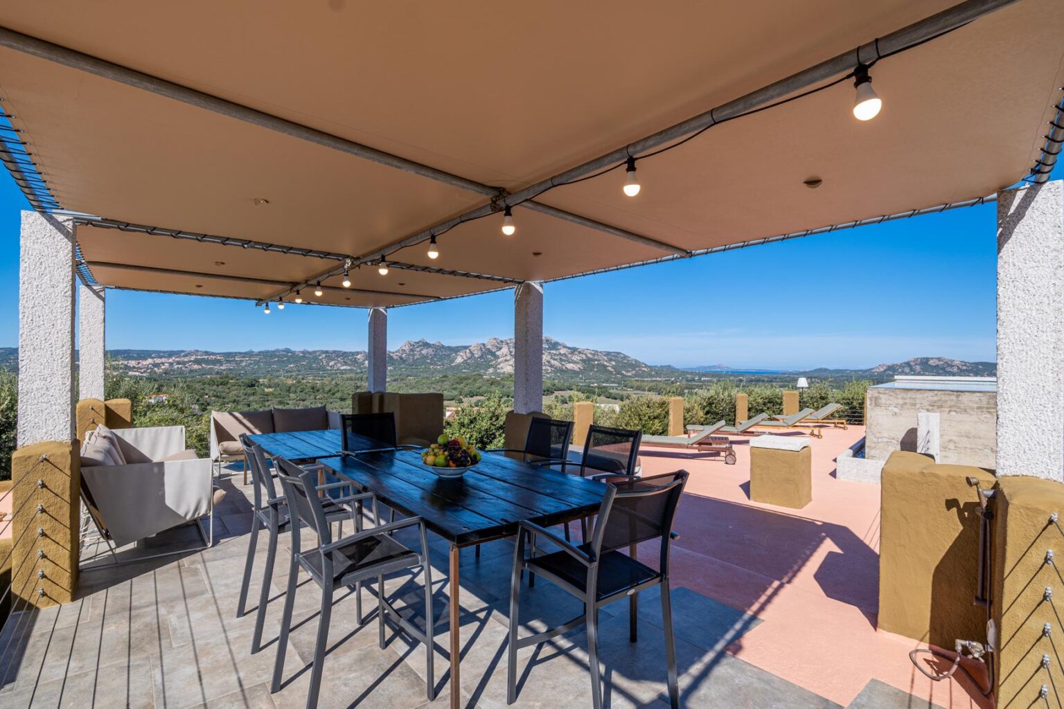Spacious outdoor dining area with a wooden table and chairs, surrounded by views of mountains and greenery, under a shaded canopy with string lights, ideal for gatherings and relaxation. | Only on SopranoVillas