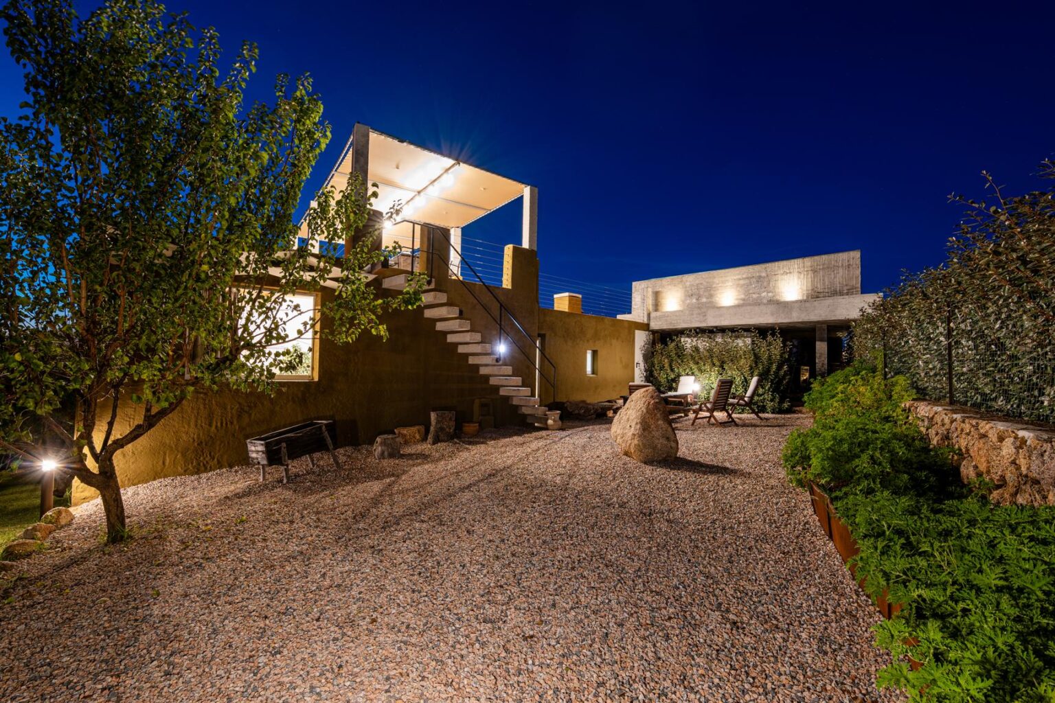 Modern architectural home with a landscaped garden at night, featuring a gravel pathway, outdoor seating, and illuminated stairs leading to a rooftop terrace. | Only on SopranoVillas