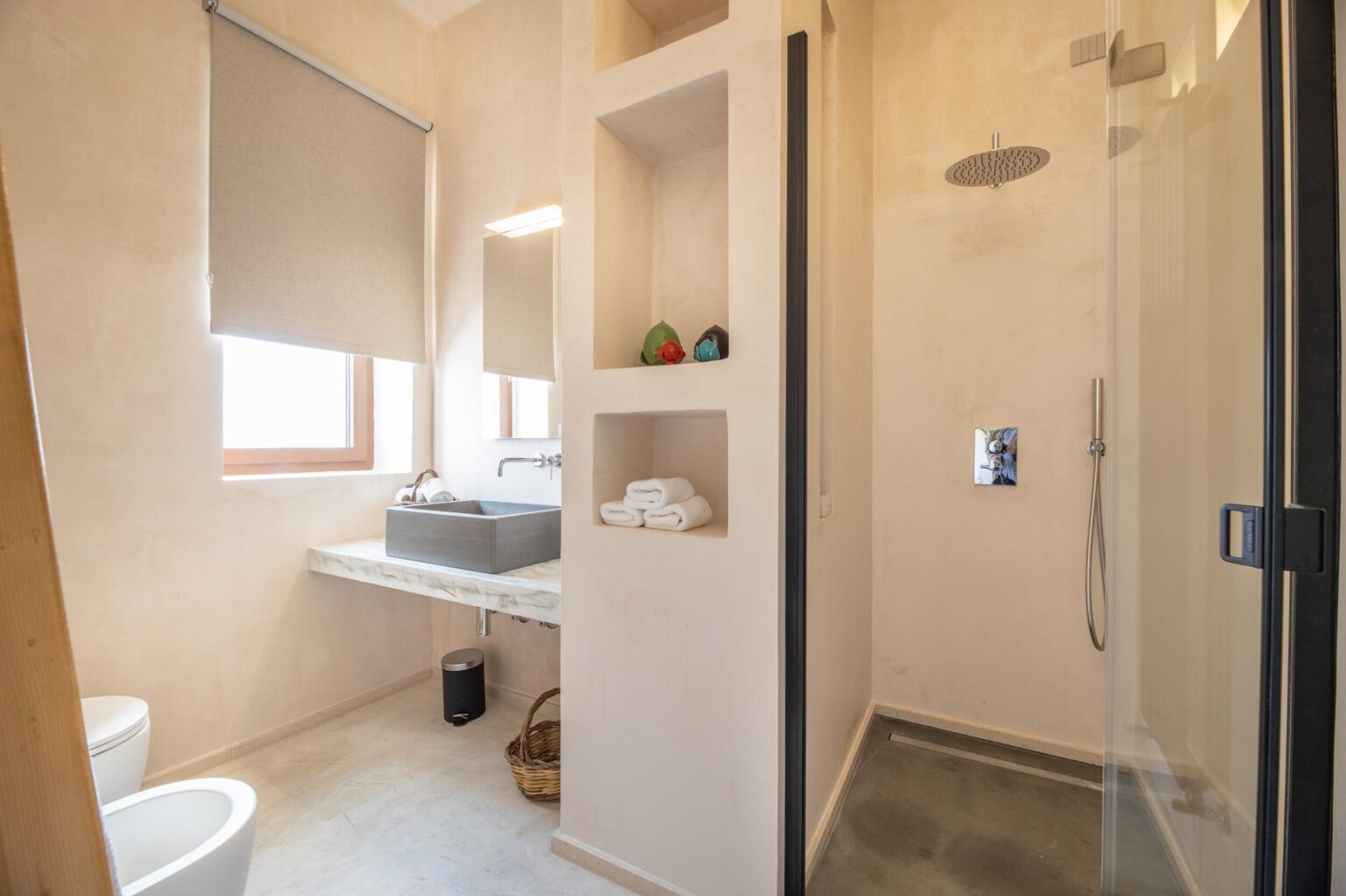 Modern bathroom featuring a glass shower enclosure, minimalist design, and natural light from a window. The space includes a sleek vanity with a sink, neatly organized toiletries, and a toilet, showcasing a clean and contemporary aesthetic. | Only on SopranoVillas