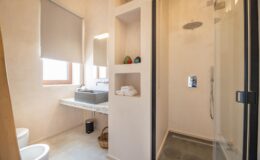 Modern bathroom featuring a glass shower enclosure, minimalist design, and natural light from a window. The space includes a sleek vanity with a sink, neatly organized toiletries, and a toilet, showcasing a clean and contemporary aesthetic.