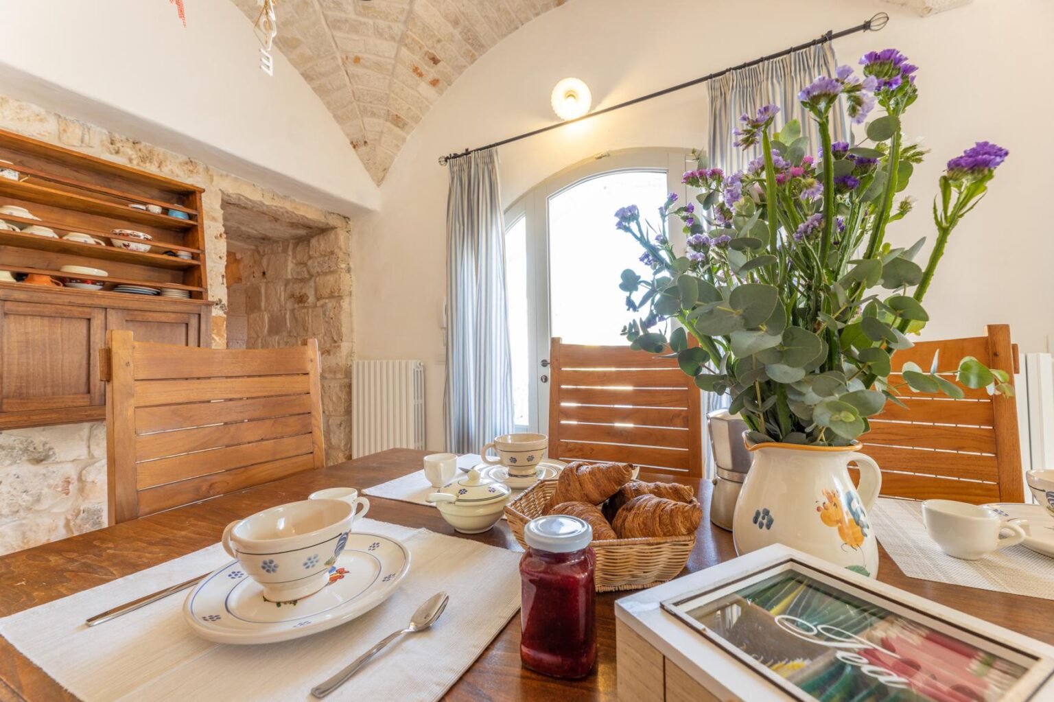 Cozy dining area featuring a wooden table set for breakfast with elegant dishware, a basket of croissants, and a jar of jam. A vibrant bouquet of flowers adds a touch of color, while stone walls and large windows create a warm, inviting atmosphere. Perfect for a relaxed morning gathering. | Only on SopranoVillas