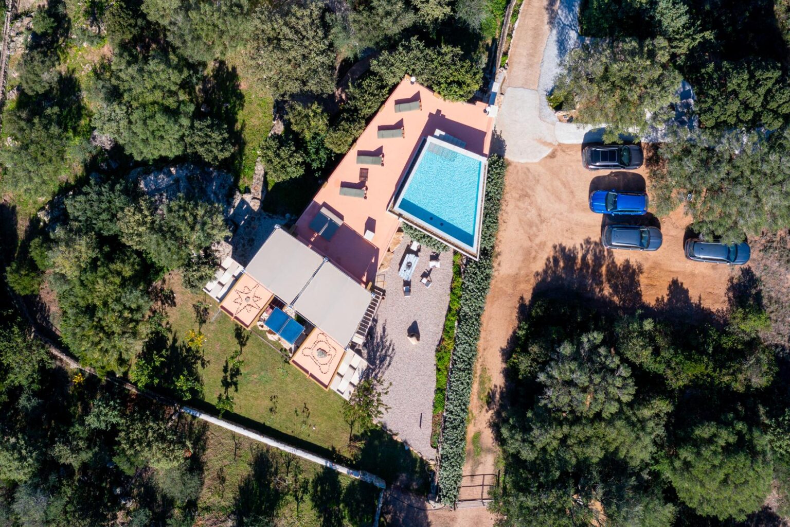 Aerial view of a modern villa surrounded by lush greenery, featuring a swimming pool, outdoor seating area, and multiple vehicles parked nearby. | Only on SopranoVillas