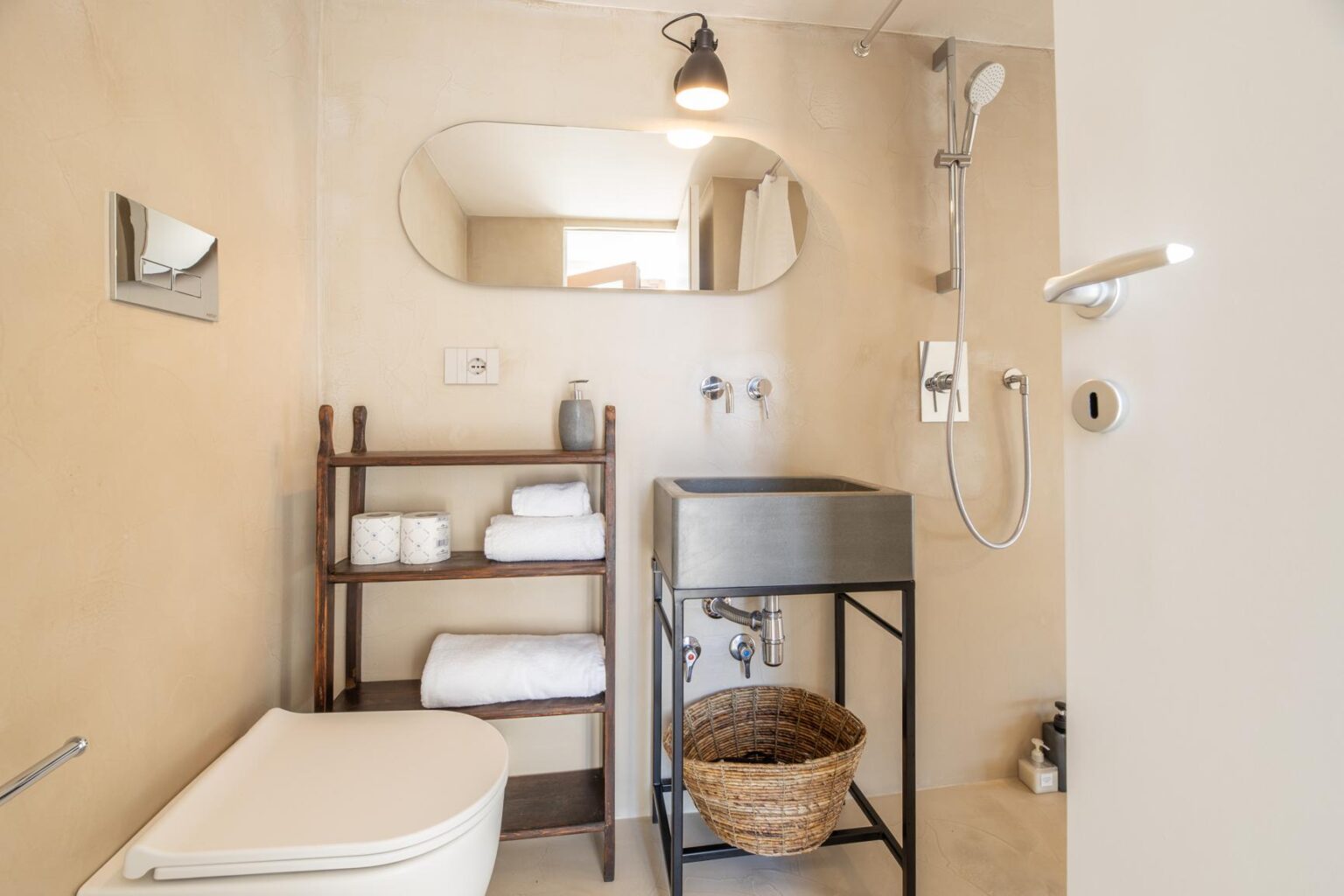 Modern bathroom featuring a shower, minimalist design, and natural light. Includes a stylish mirror, wooden shelving with neatly stacked towels, and a decorative basket, creating a serene and functional space. | Only on SopranoVillas