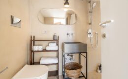 Modern bathroom featuring a shower, minimalist design, and natural light. Includes a stylish mirror, wooden shelving with neatly stacked towels, and a decorative basket, creating a serene and functional space.