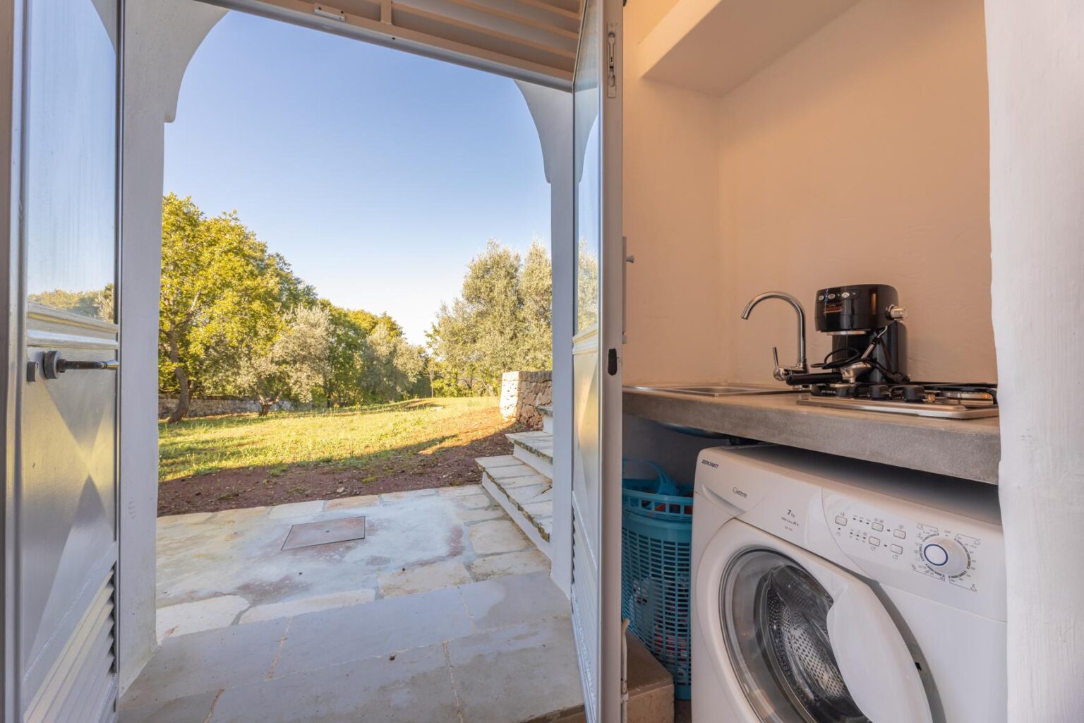 Laundry room with a washing machine and sink, opening to a scenic outdoor view of greenery and trees. | Only on SopranoVillas