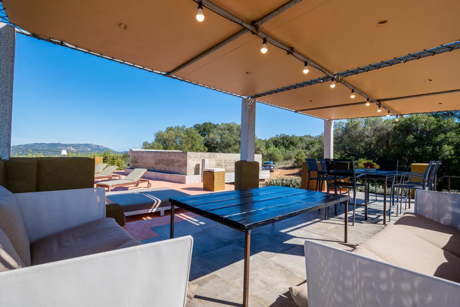 Outdoor patio area with comfortable seating, dining tables, and lounge chairs under a shaded canopy, surrounded by greenery and mountains in the background. Ideal for relaxation and gatherings. | Only on SopranoVillas