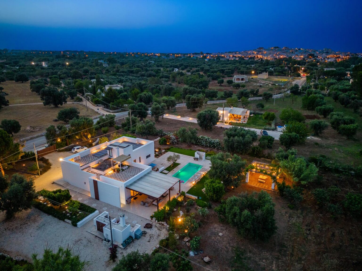 Aerial view of a modern villa with a swimming pool, surrounded by olive trees and lush greenery, illuminated at dusk, showcasing the serene landscape and nearby town in the background. | Only on SopranoVillas