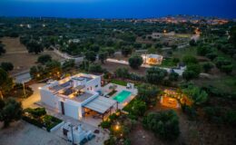 Aerial view of a modern villa with a swimming pool, surrounded by olive trees and lush greenery, illuminated at dusk, showcasing the serene landscape and nearby town in the background.