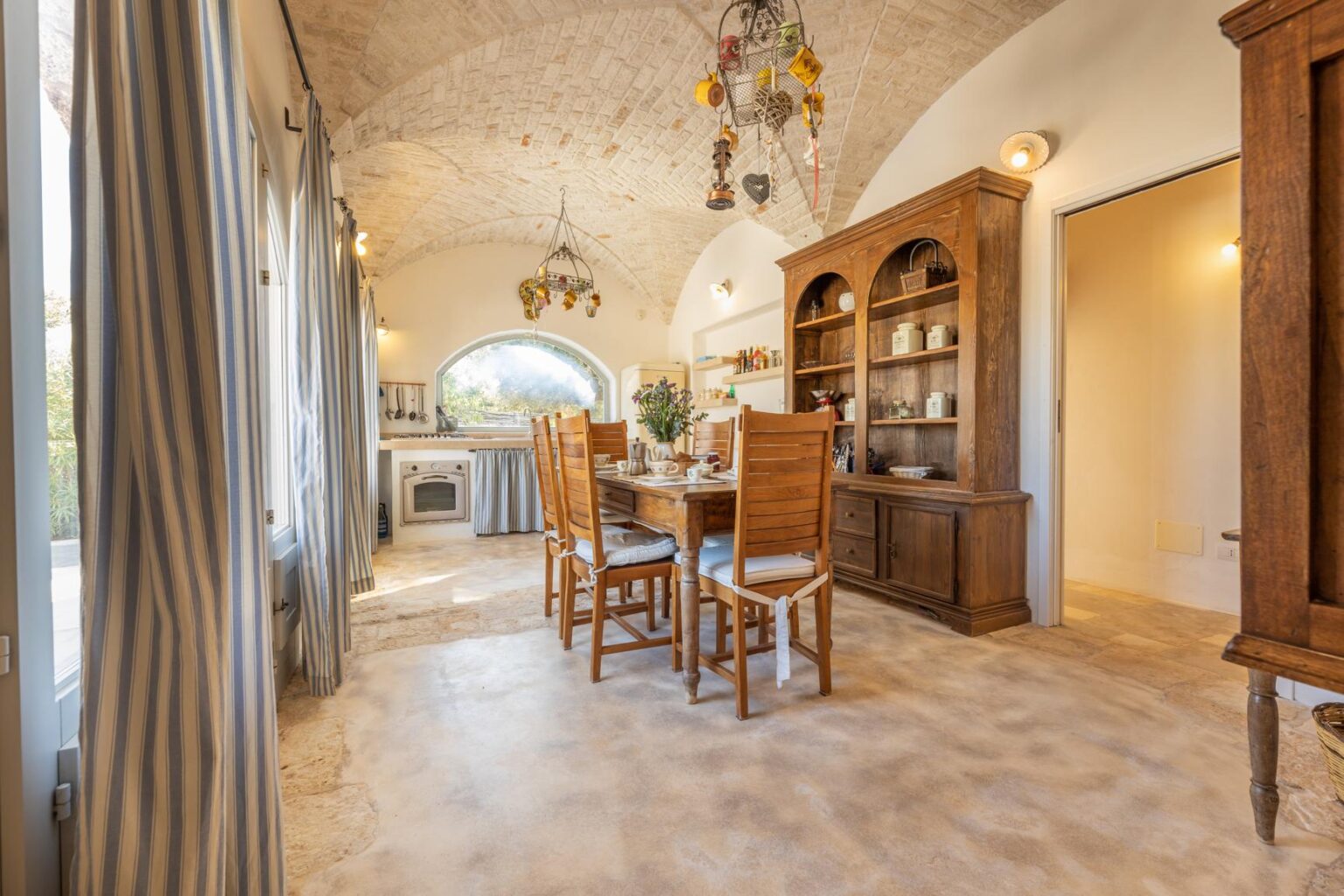 Cozy dining area featuring a wooden dining table with chairs, a rustic cabinet, and large windows allowing natural light, set in a stylishly decorated room with arched ceilings. | Only on SopranoVillas