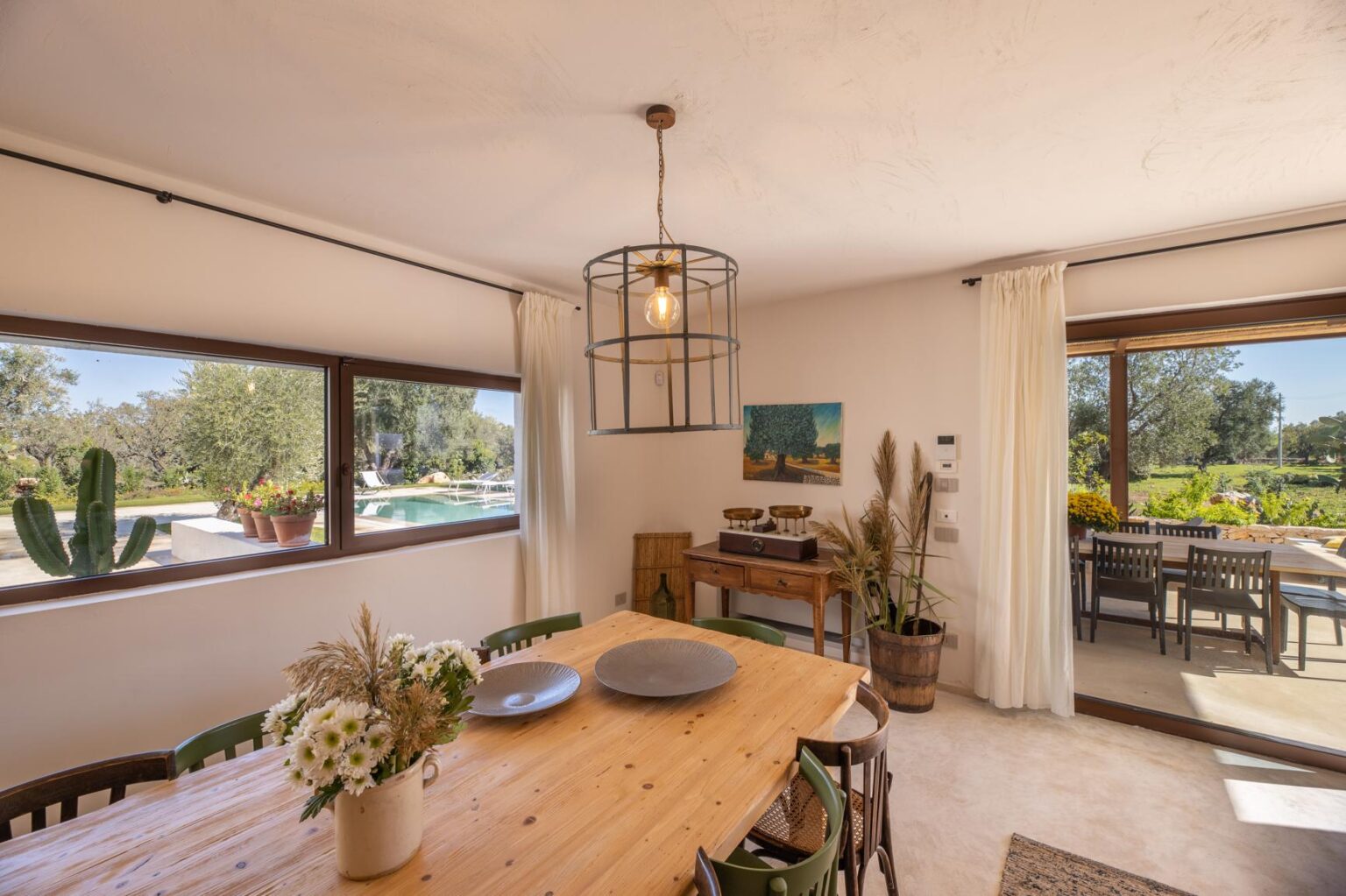 Bright and airy dining area featuring a wooden table, stylish pendant light, and large windows overlooking a serene outdoor pool and garden. The space is decorated with natural elements and a cozy aesthetic, perfect for entertaining. | Only on SopranoVillas