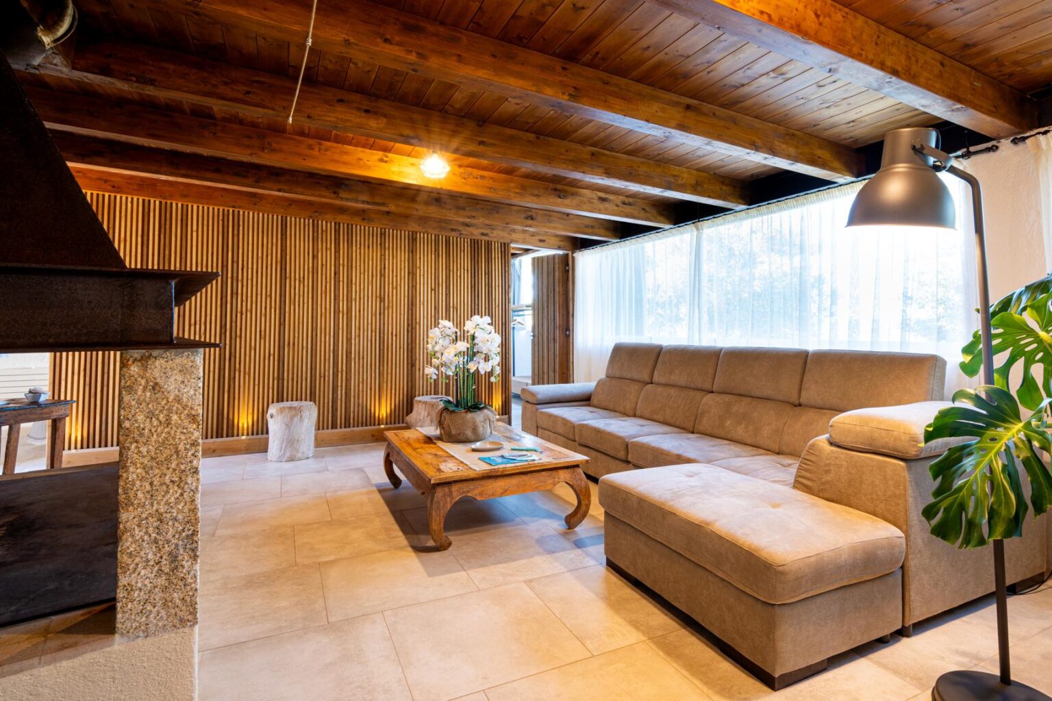 Cozy modern living room featuring a beige sectional sofa, wooden ceiling beams, and large windows with sheer curtains. A decorative coffee table sits on tiled flooring, complemented by indoor plants and elegant lighting. | Only on SopranoVillas