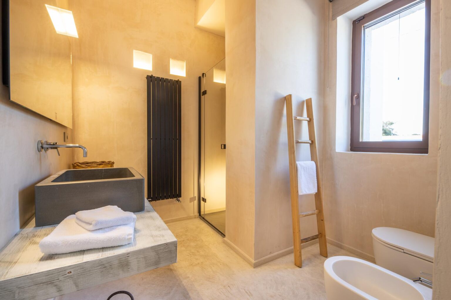 Modern bathroom featuring a minimalist design with a concrete sink, walk-in shower, and natural light from a window. Towel rack and neatly arranged towels enhance the aesthetic appeal. | Only on SopranoVillas