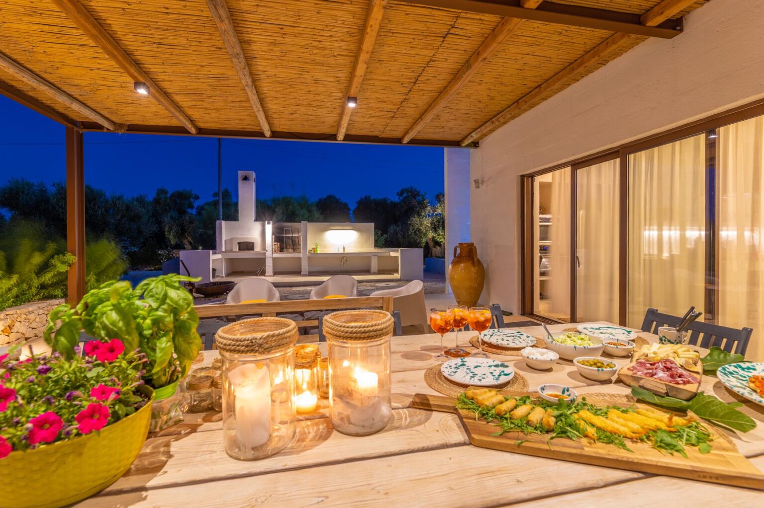 Outdoor dining area at night featuring a beautifully arranged table with gourmet dishes, candles, and fresh herbs, surrounded by lush greenery and a modern outdoor kitchen in the background. | Only on SopranoVillas