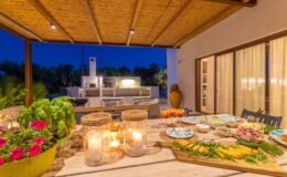 Outdoor dining area at night featuring a beautifully arranged table with gourmet dishes, candles, and fresh herbs, surrounded by lush greenery and a modern outdoor kitchen in the background.
