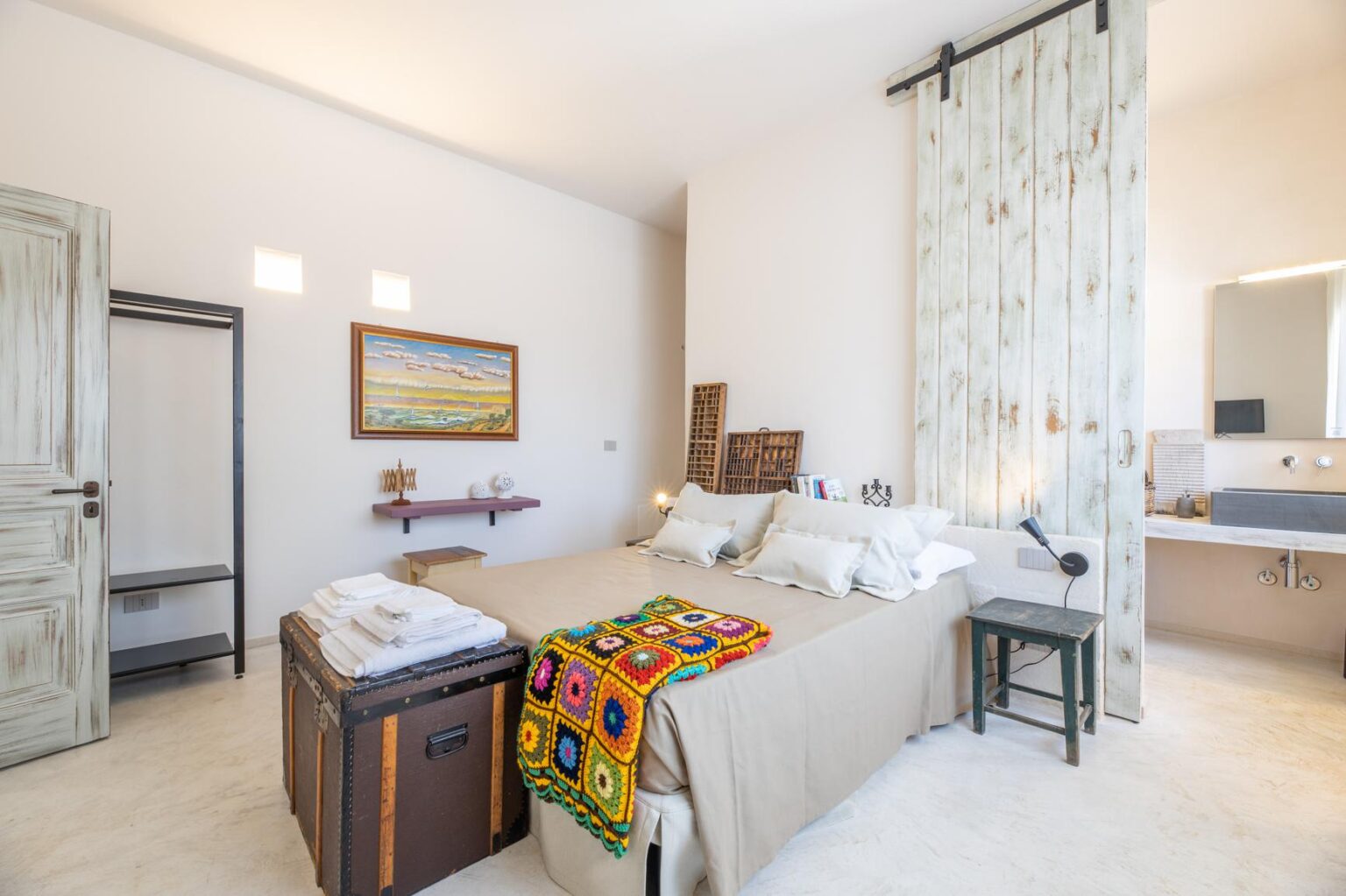 Cozy and modern bedroom featuring a queen-sized bed with decorative pillows, a colorful crochet blanket, and a vintage trunk. The room includes a rustic sliding barn door, a minimalist closet, and a bathroom area with a stylish sink and mirror. Natural light floods the space, complemented by artwork and decorative elements. | Only on SopranoVillas