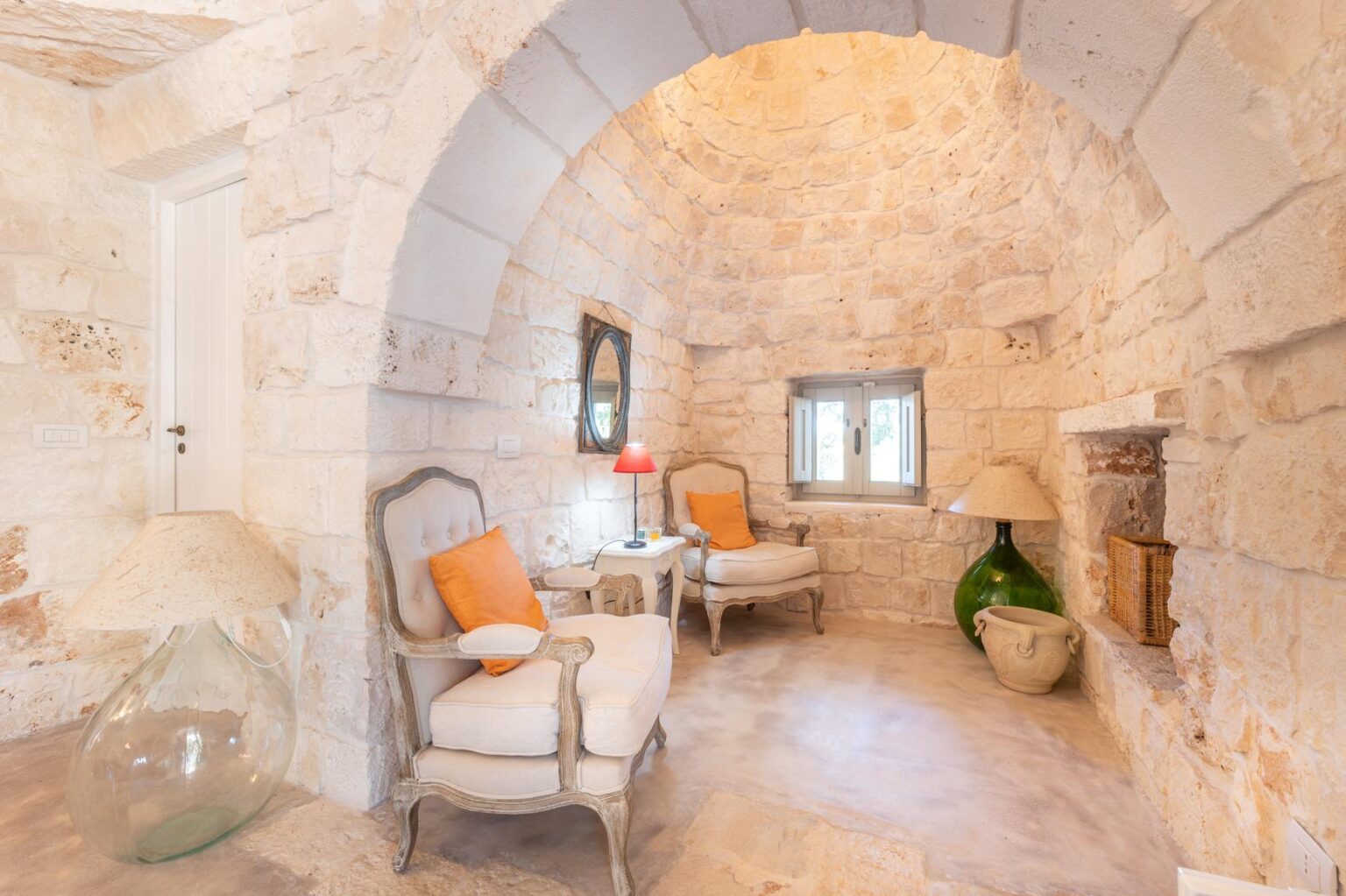 Cozy interior of a stone-walled living space featuring two armchairs with orange cushions, a decorative lamp, and a large green vase, illuminated by natural light from a window. | Only on SopranoVillas
