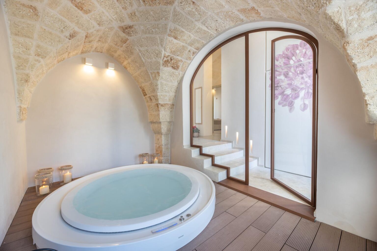 A modern spa room featuring a circular hot tub, elegant stone archways, and soft lighting. The space includes wooden flooring and a staircase leading to a serene area with floral decor. Perfect for relaxation and wellness retreats. | Only on SopranoVillas