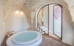 A modern spa room featuring a circular hot tub, elegant stone archways, and soft lighting. The space includes wooden flooring and a staircase leading to a serene area with floral decor. Perfect for relaxation and wellness retreats.