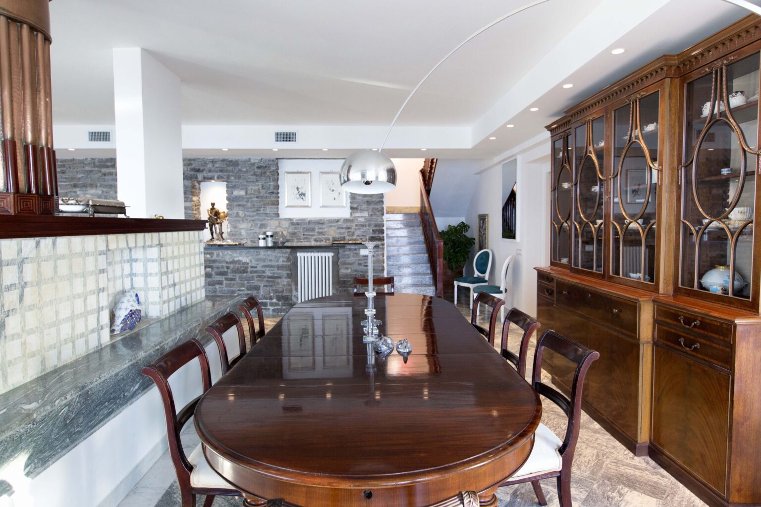 Elegant dining room featuring a large wooden table surrounded by chairs, a stylish lamp overhead, and a decorative sideboard with glass display. The room showcases a stone accent wall and modern decor, creating a sophisticated atmosphere. | Only on SopranoVillas