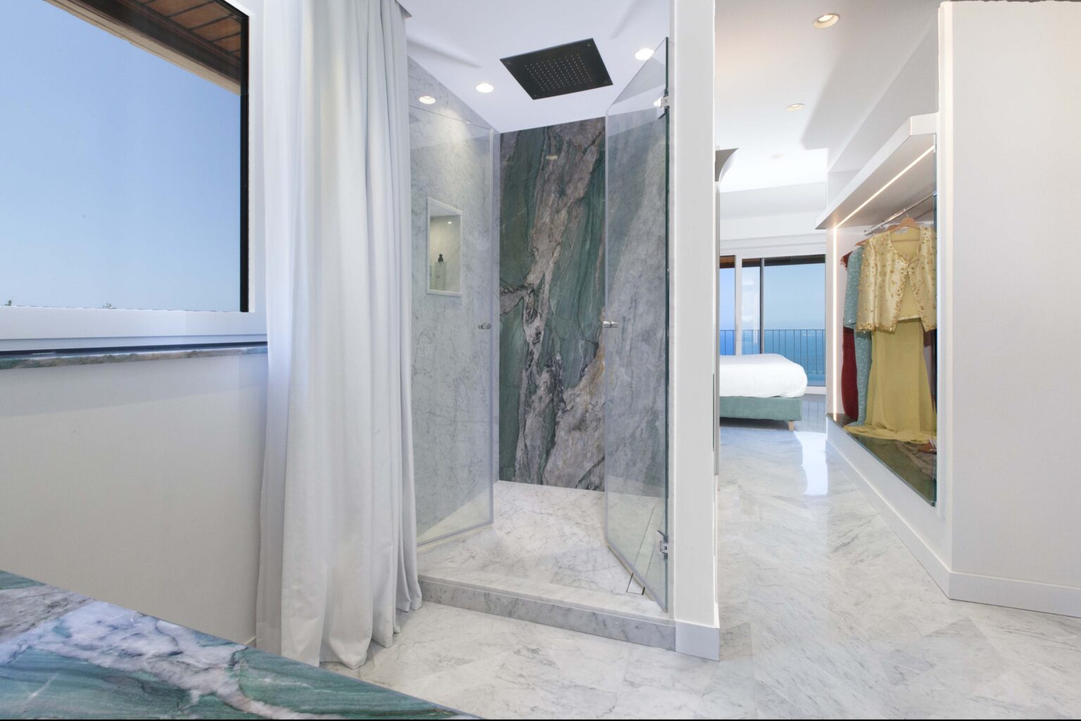Modern bathroom featuring a glass shower with green and gray marble walls, a sleek design, and a view of the ocean through a large window. Elegant dressing area with hanging garments is visible in the background. | Only on SopranoVillas