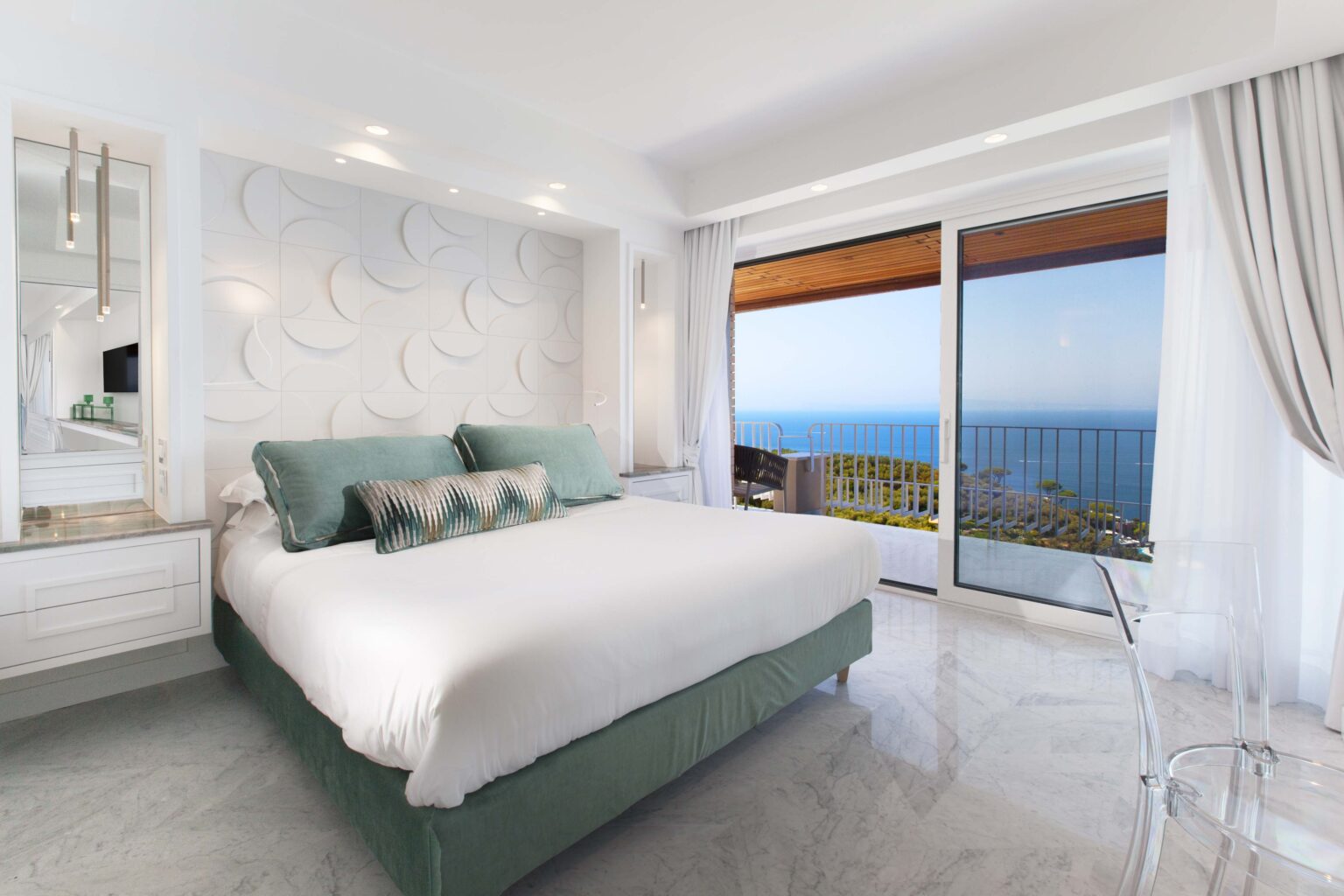 Modern hotel bedroom featuring a plush king-size bed with decorative pillows, sleek white walls, and large glass doors opening to a stunning ocean view. The room includes stylish furnishings, marble flooring, and contemporary lighting, creating a serene and luxurious atmosphere. | Only on SopranoVillas