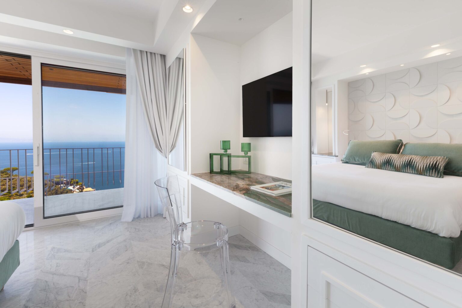 Modern hotel room with a stunning ocean view, featuring a comfortable bed with decorative pillows, a sleek desk with a transparent chair, and a large flat-screen TV mounted on the wall. The room is adorned with light-colored marble flooring and elegant curtains, creating a bright and inviting atmosphere. | Only on SopranoVillas