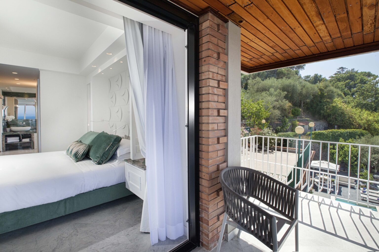 Modern bedroom with green accent pillows, featuring a balcony view of lush greenery and outdoor seating. The interior showcases sleek design elements and ample natural light. Perfect for a relaxing getaway. | Only on SopranoVillas