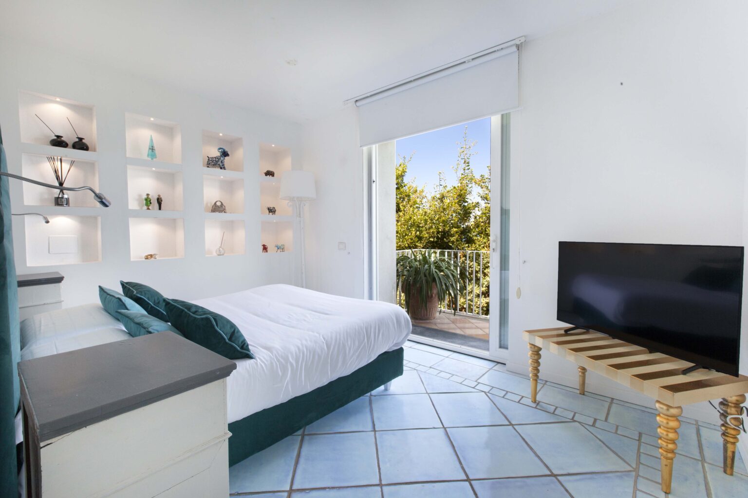 Bright and airy bedroom featuring a queen-sized bed with green accent pillows, a flat-screen TV, and a balcony door leading to a scenic outdoor view. The room has modern shelving with decorative items and a minimalist design. | Only on SopranoVillas