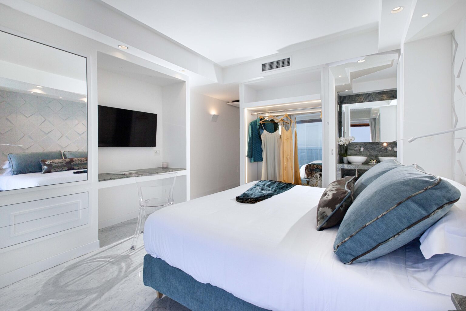 Modern hotel bedroom featuring a plush king-sized bed with decorative pillows, a flat-screen TV, stylish mirrors, and a spacious closet. The room is well-lit with natural light, showcasing elegant decor and a serene color palette. | Only on SopranoVillas