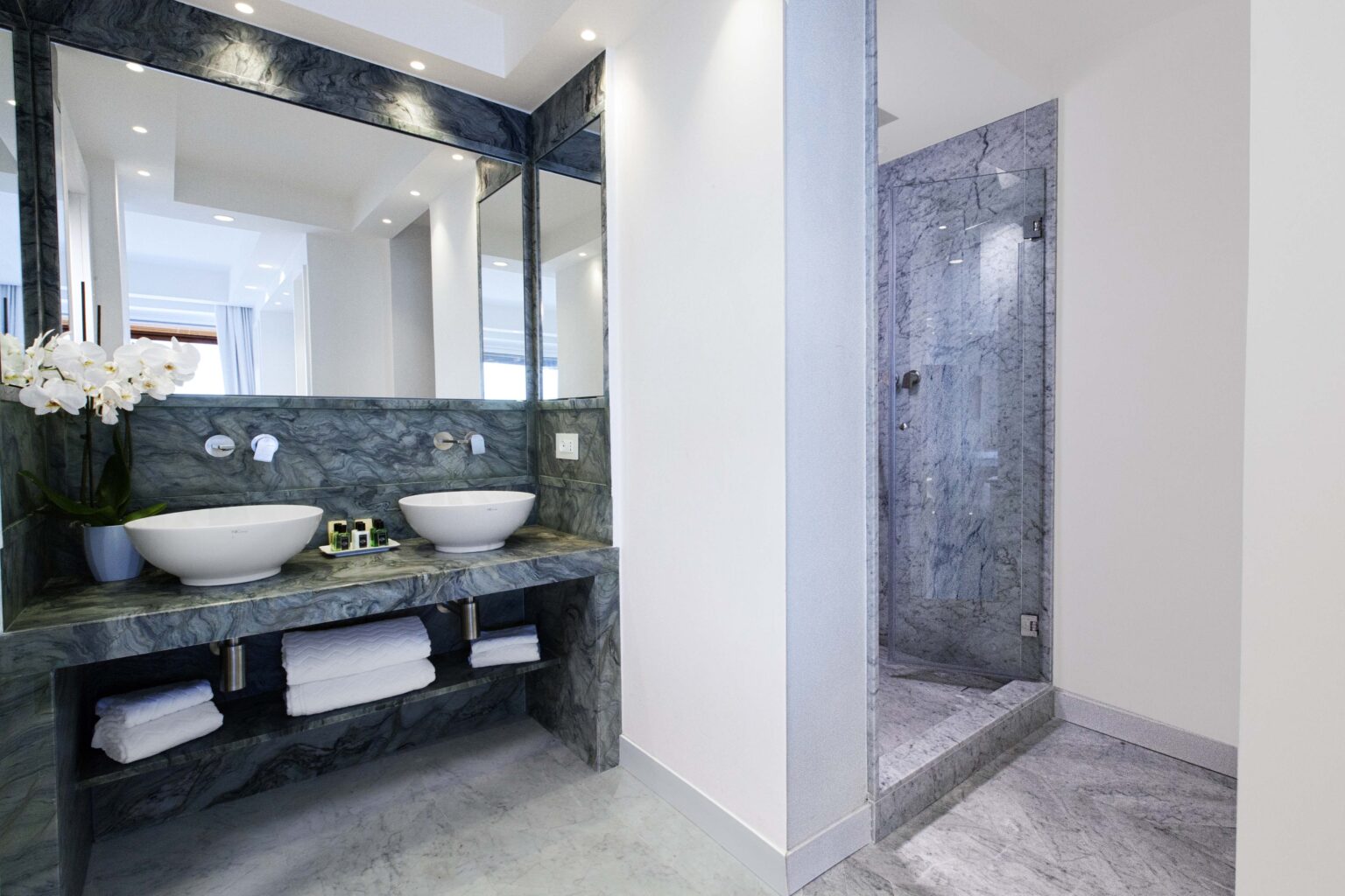 Modern bathroom featuring a sleek marble vanity with two sinks, a large mirror, and a glass shower stall. Elegant decor includes fresh flowers and neatly stacked towels, creating a luxurious and stylish atmosphere. | Only on SopranoVillas