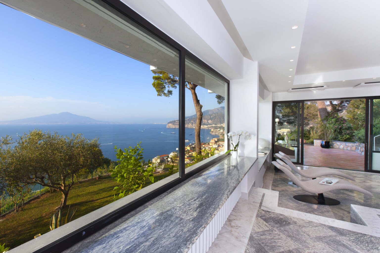 Modern interior with large windows showcasing a stunning view of the sea and mountains, featuring a sleek marble countertop and contemporary furniture. | Only on SopranoVillas