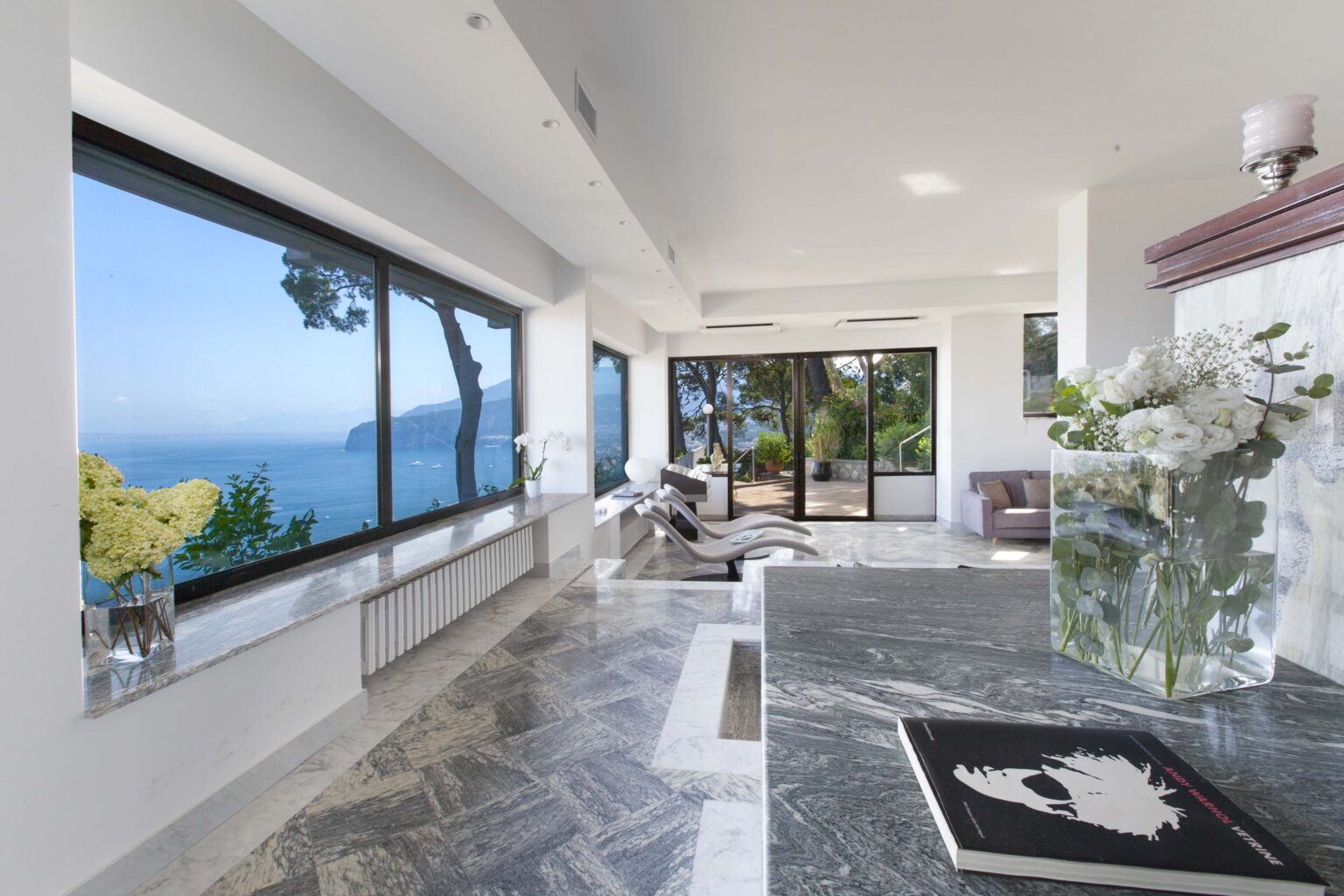 Modern living room with panoramic ocean views, featuring sleek furniture, marble flooring, and a stylish vase of white flowers on a granite countertop. The large windows allow natural light to flood the space, creating an inviting atmosphere. | Only on SopranoVillas