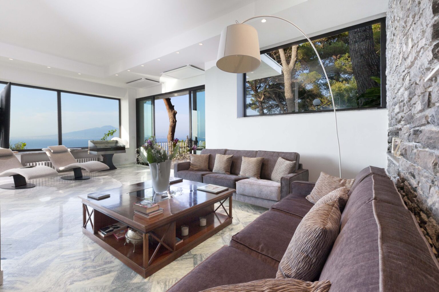 Modern living room with large windows offering ocean views, featuring a comfortable sofa, stylish coffee table, and contemporary lighting. Elegant decor and natural light create a serene atmosphere. | Only on SopranoVillas