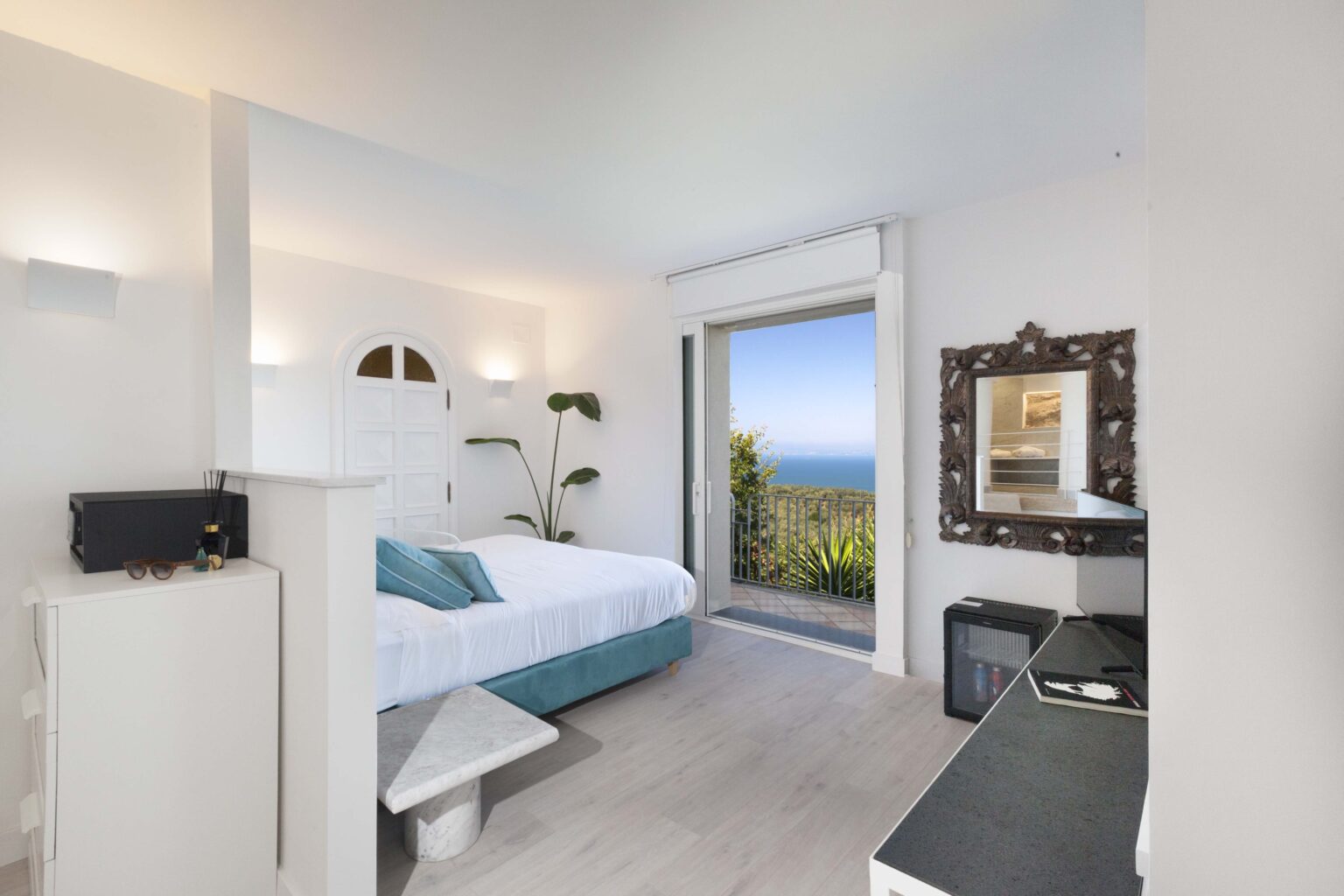 Modern hotel room with a queen bed, featuring a coastal view through large windows, minimalist decor, and a stylish mirror. Ideal for a relaxing getaway. | Only on SopranoVillas