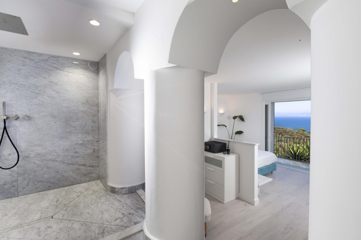 Modern bathroom with a spacious walk-in shower featuring marble walls, leading to a bright bedroom with ocean views. The design includes elegant arches and natural light, creating a serene atmosphere. | Only on SopranoVillas