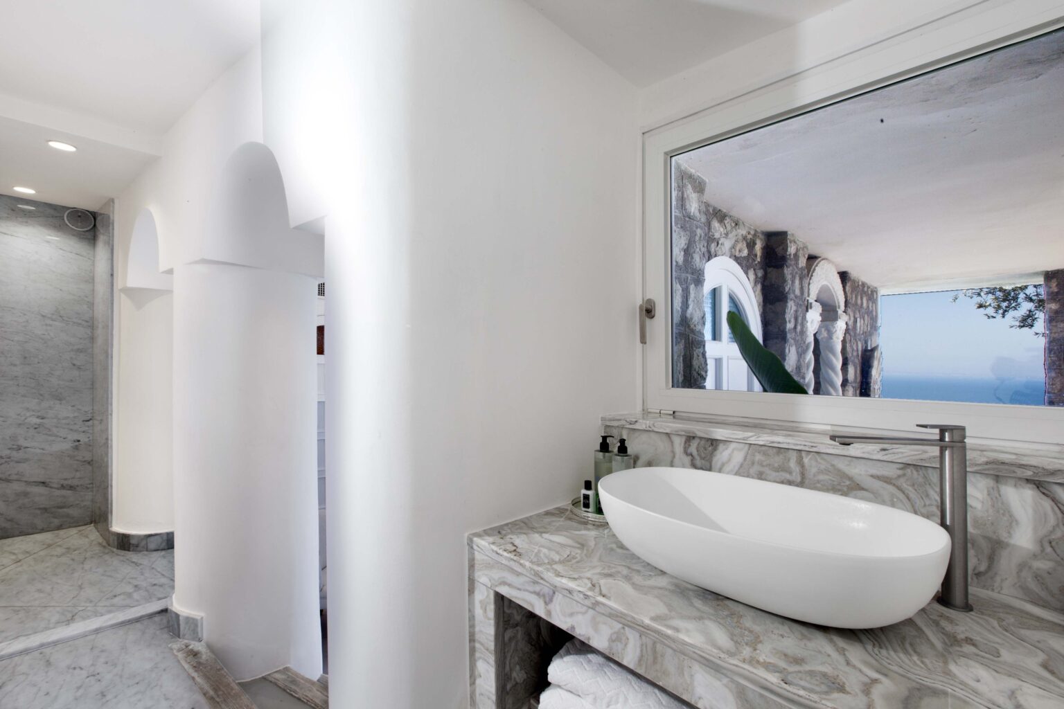 Modern bathroom interior featuring a sleek white vessel sink on a marble countertop, a spacious shower area, and a large window offering a scenic view. The design emphasizes clean lines and a minimalist aesthetic. | Only on SopranoVillas