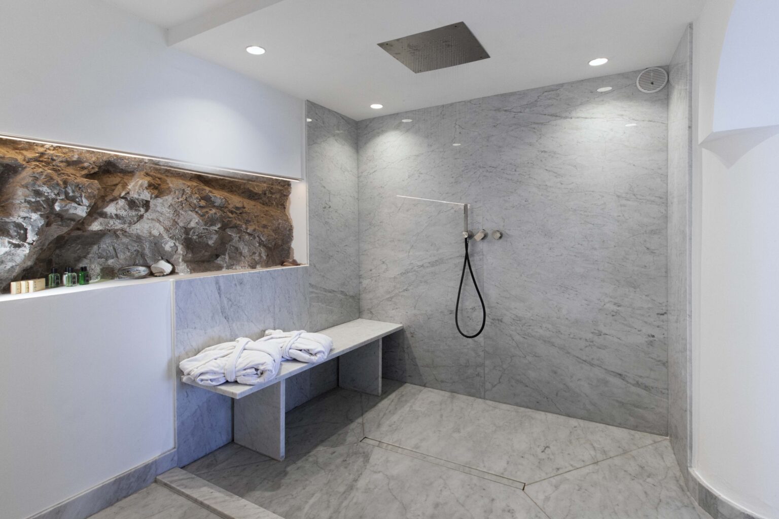 Modern bathroom with a sleek marble shower wall, minimalist design, and a built-in stone feature. Features a showerhead, bench seating, and neatly arranged towels for a spa-like ambiance. | Only on SopranoVillas