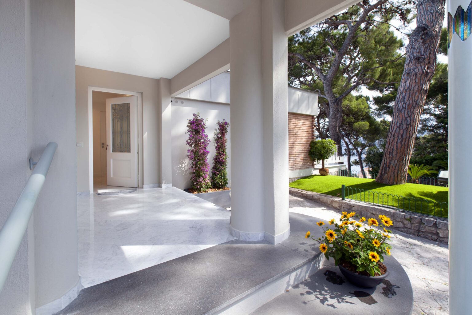 Spacious and inviting entryway with modern architecture, featuring a polished stone floor, large windows, and vibrant flower beds, surrounded by lush greenery. | Only on SopranoVillas