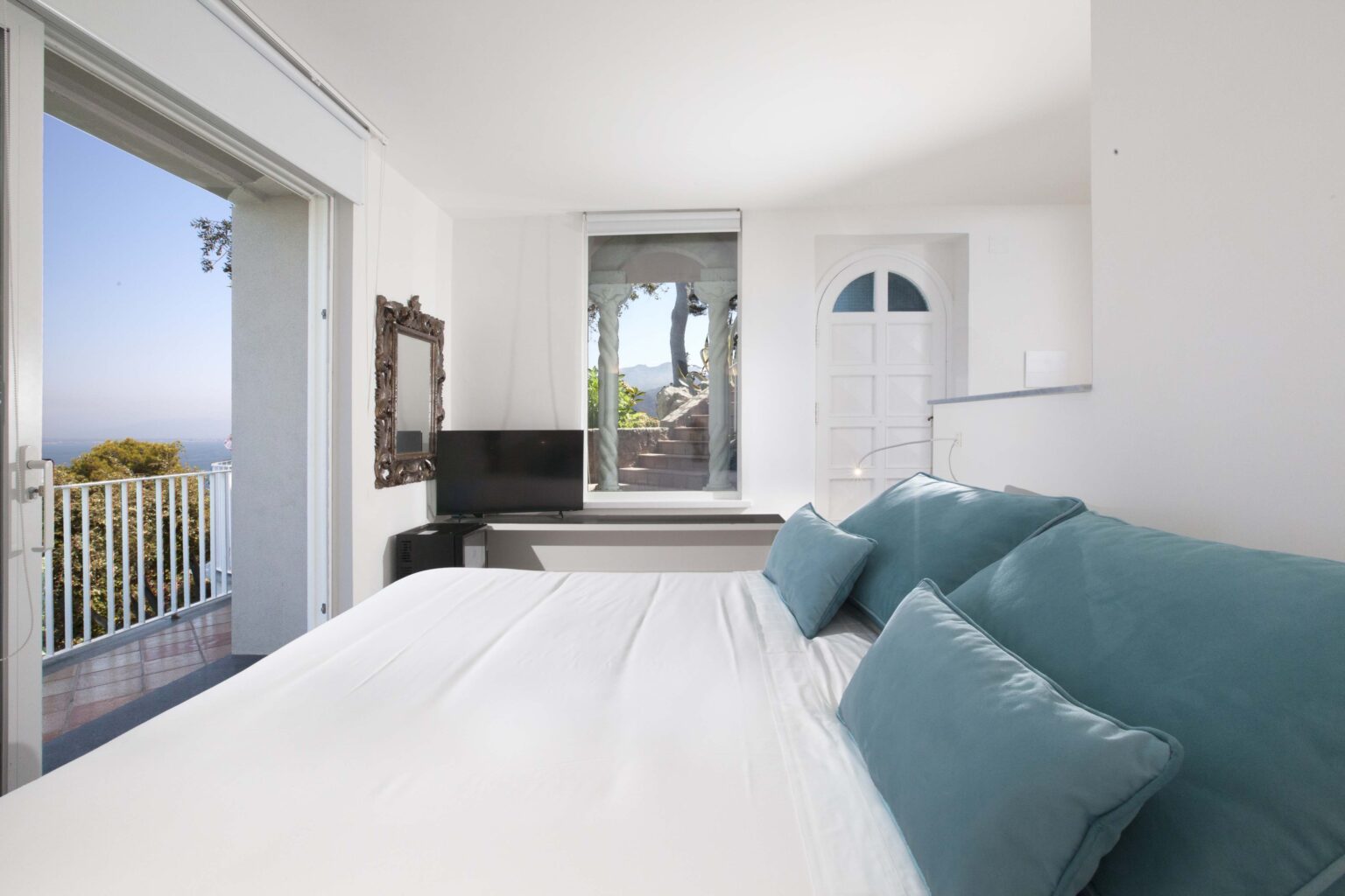 Bright and airy bedroom featuring a large bed with blue cushions, a flat-screen TV, and a balcony with scenic views. The room has white walls and modern decor, creating a serene atmosphere. | Only on SopranoVillas