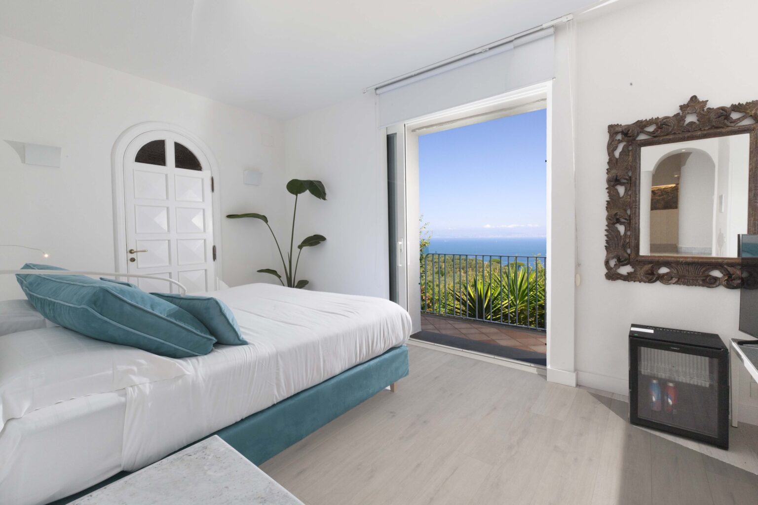 Modern bedroom with a large window showcasing a stunning ocean view, featuring a comfortable bed with teal accents, minimalist decor, and a mirror. | Only on SopranoVillas
