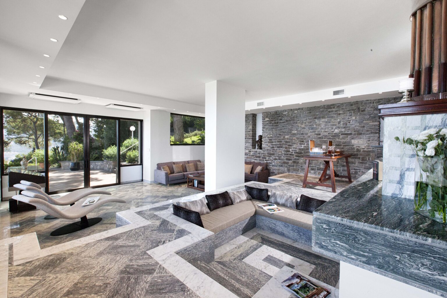 Modern interior of a stylish living room featuring elegant stone walls, a unique sunken seating area, and large glass windows that offer a view of the lush outdoor landscape. The space includes contemporary furniture, decorative items, and a sophisticated bar setup, creating a luxurious atmosphere. | Only on SopranoVillas